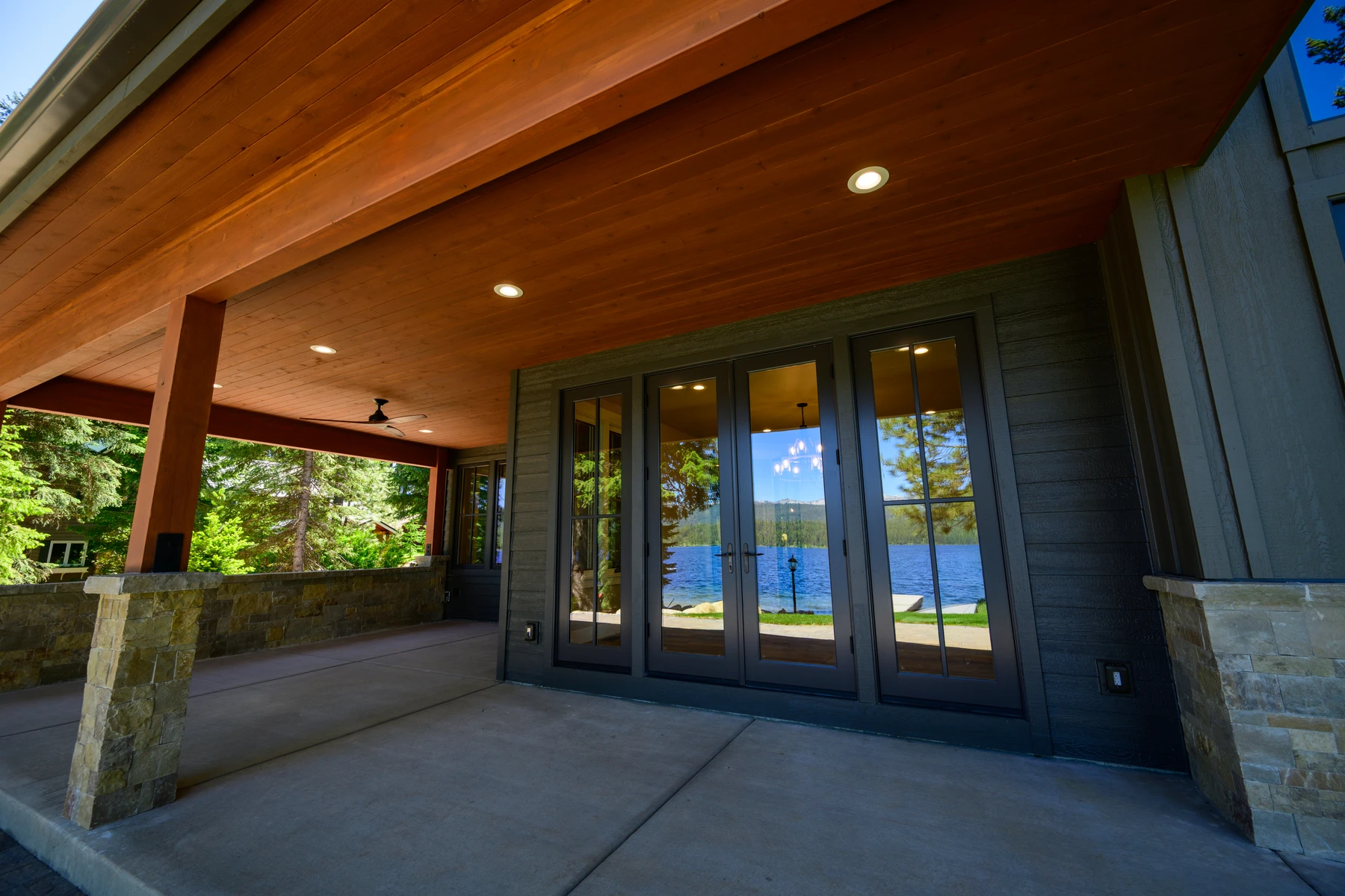 Covered lakefront patio with a wood ceiling, stone columns, and glass doors
